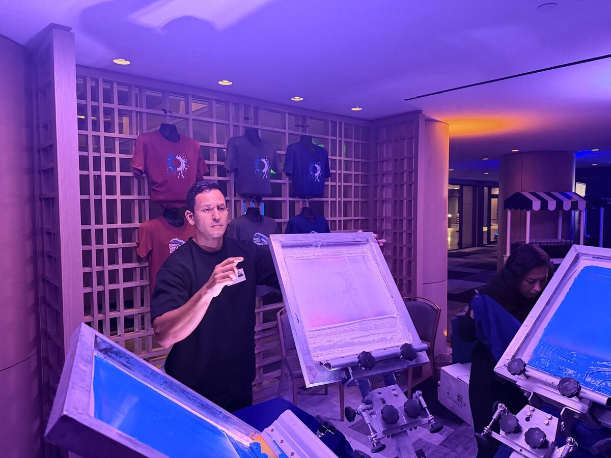 Operator preparing a live tee printing station beside a wall of display shirts at an evening reception